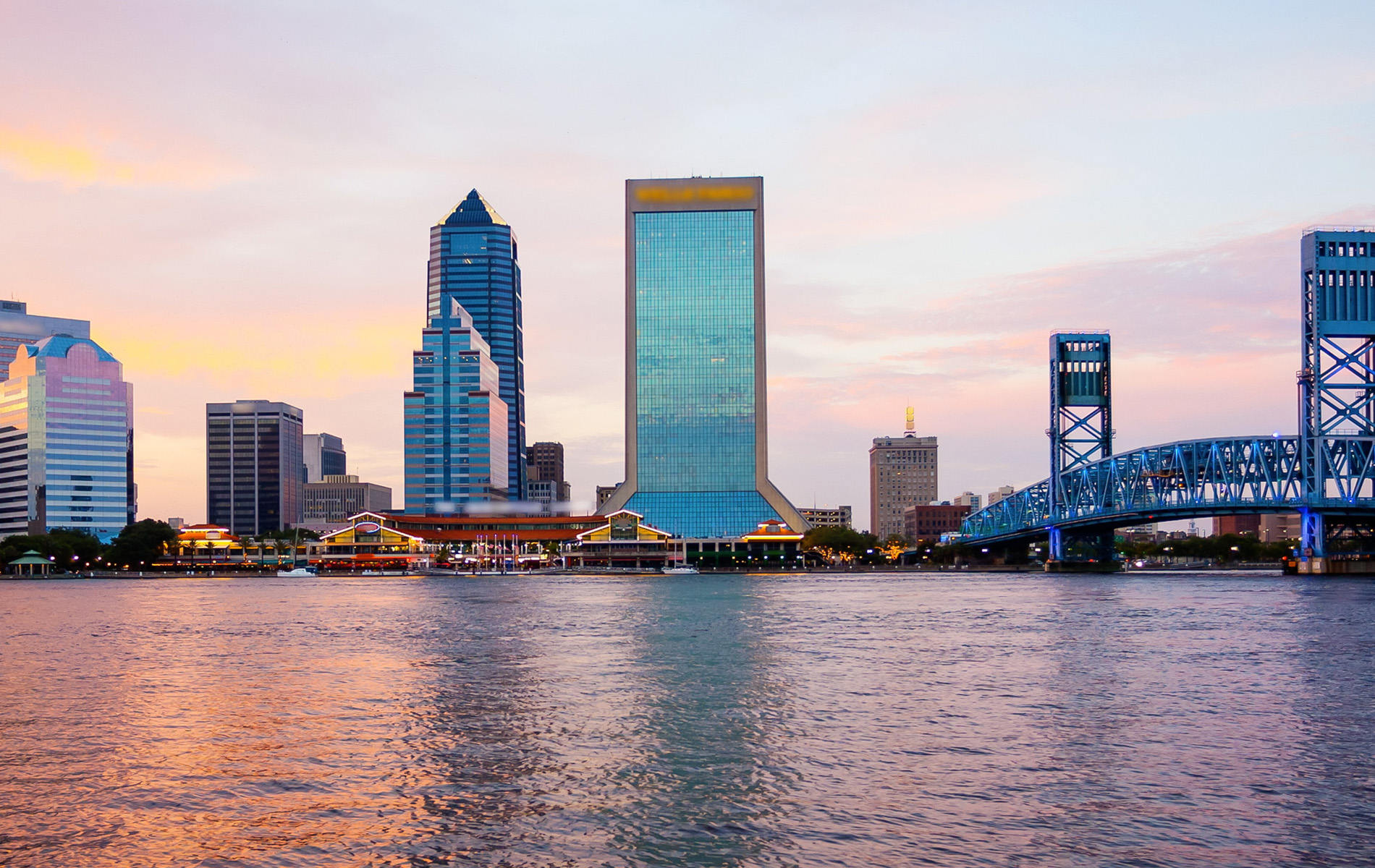 A city skyline at dusk, featuring a prominent skyscraper with a reflective facade and the silhouette of a bridge against a colorful sunset.