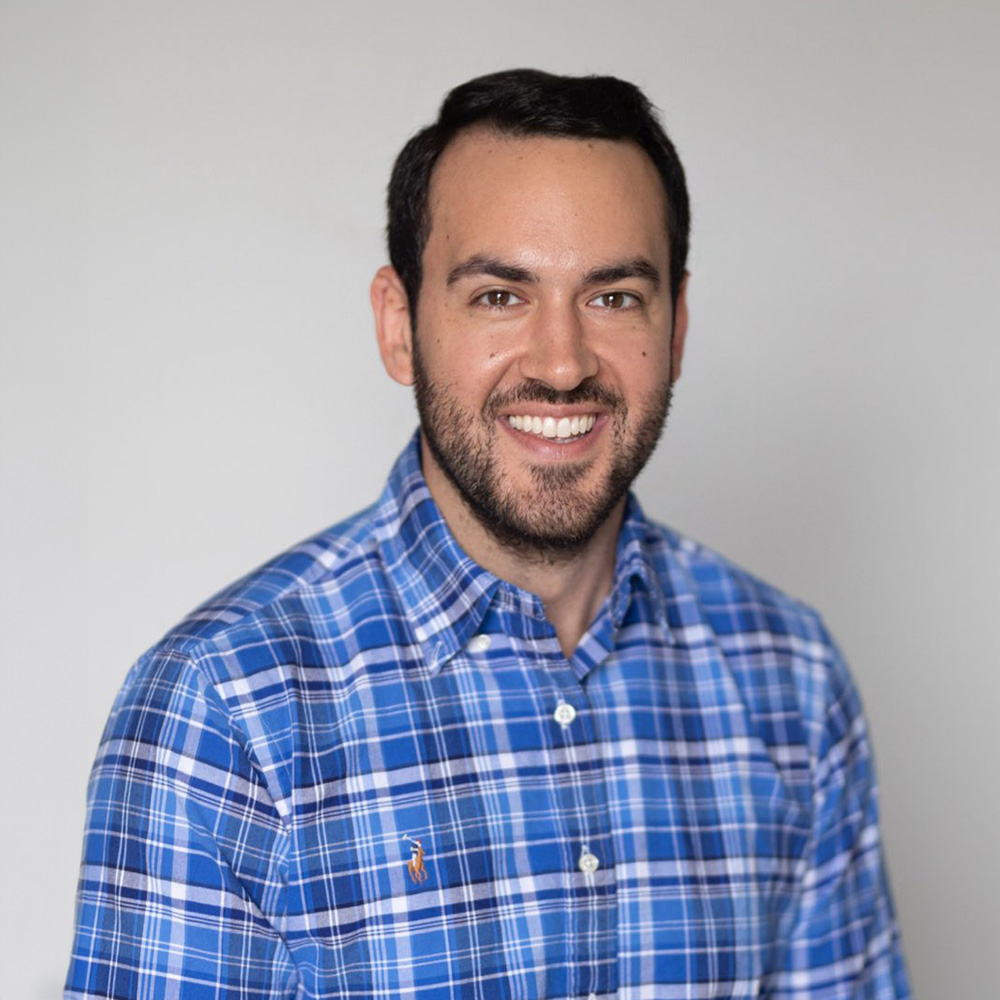 The image shows a man with a beard smiling at the camera. He is wearing a blue plaid shirt and has short hair. The background is plain and does not provide any additional context about the location or setting of the photo.