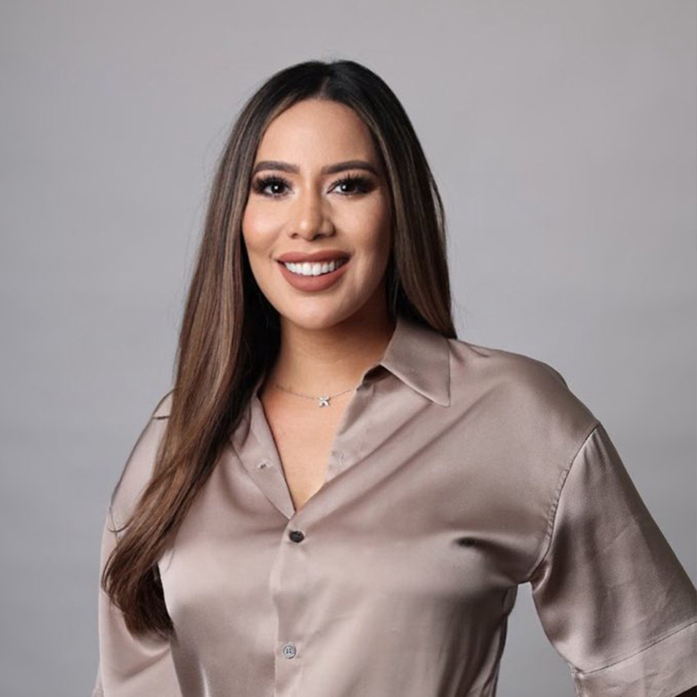 A woman with long dark hair, wearing a light brown blazer and smiling.