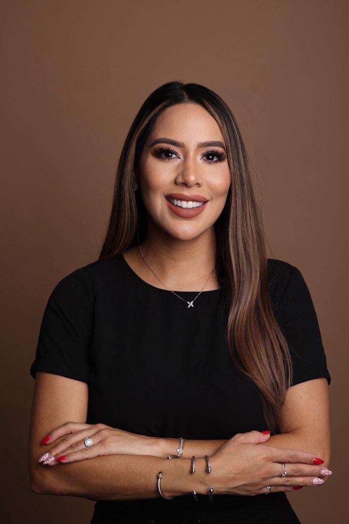 The image features a woman with long dark hair, wearing a black top and posing in front of a brown background. She is smiling and has her arms crossed over her chest.
